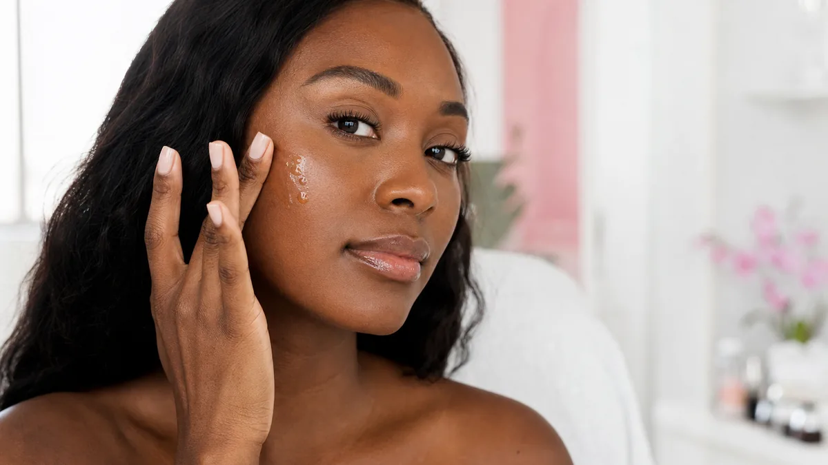 Woman with a deeper skin tone applying vitamin C serum to her cheek for a brighter, more even tone
