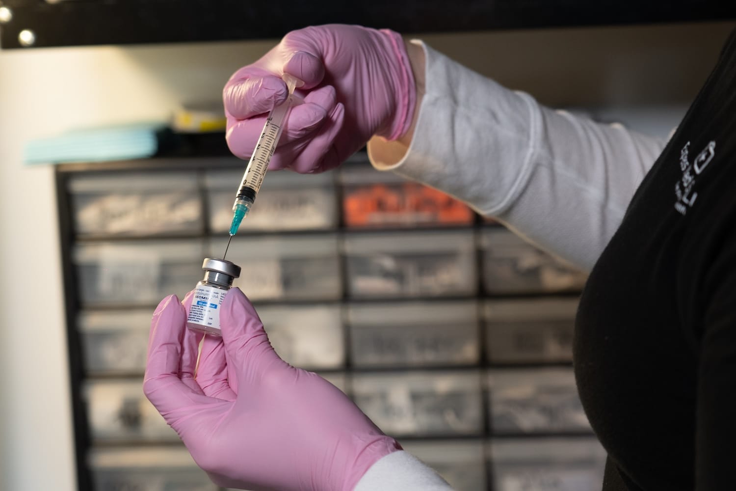 Pink-gloved hands drawing liquid from a small vial into a syringe in front of a wall of storage drawers